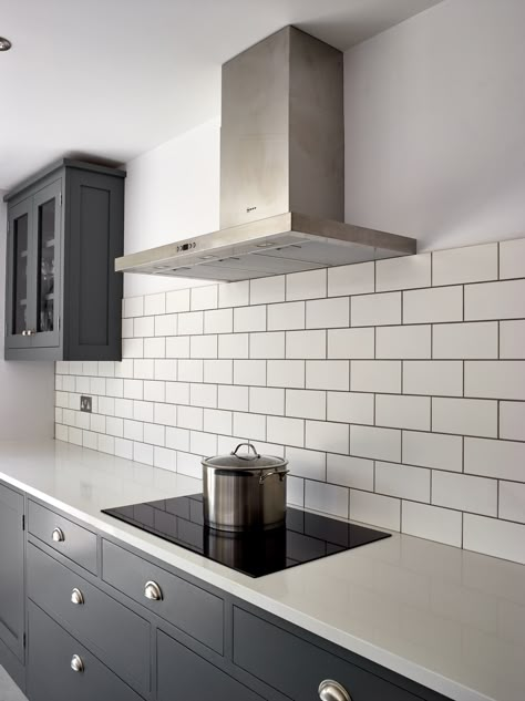 The white metro tiles create nice contrast with the Shaker kitchen cupboards painted in a dark grey colour, Little Greene 'Scree'. The worktops are Caesarstone 'Snow' in a polished finish and a 30mm thickness whilst the induction hob is T58FD20X0 from Neff. Also the freestanding stainless steel extractor is by Neff. The cup handles are from the Cotswold range by Armac Martin. Kitchen Tiles Grey Kitchen, Dark Grey Kitchen White Worktop, Metro Tiles Kitchen White, Grey Kitchen White Tiles, Grey Kitchen Tiles Wall, White Worktop Kitchen, Tile Paint Kitchen, Gold Kitchen Decor Ideas, Symmetrical Kitchen