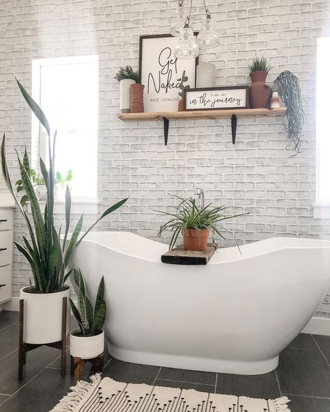 This bathroom with whitewashed brick walls features a white freestanding oval soaking tub with rustic wood bath tray. A light wood and black metal shelf is mounted above the bathtub between two white windows. Soaking Tub Decor, White Brick Bathroom, Brick Bathroom Wall, Small Bathroom Look Bigger, Get Naked Sign, Black Bathroom Floor, Wood Bath Tray, White Subway Tile Shower, Brick Bathroom