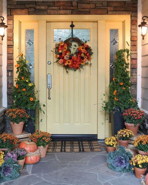 A creamy yellow door sets the stage for a warm and inviting entrance, while the plaid door mat adds a touch of charm. Standing tall on either side of the door, two potted trees lend a touch of grace to the scene. Surrounding the area, a delightful collection of potted mums in shades of yellow, gold, and orange, along with pumpkins of all sizes and textures and purple kale creates a captivating display. Learn how to decorate your fall porch this season on a budget Diy Outdoor Fall Decor, Floor Lanterns, Evergreen House, Potted Mums, Outdoor Fall Decor Ideas, Fall Front Door Decor, Wood Exterior Door, Door Paint Colors, Fall Front Door