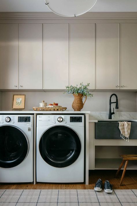 A row of light taupe cabinets are positioned over a white front loading washer and dryer enclosed beneath a marble countertop and beside a black apron sink with a matte black gooseneck faucet. Laundry Room Appliances, Transitional Laundry Room, Laundry Room Countertop, Herringbone Tile Floors, Laundry Chute, Quartz Backsplash, Checkerboard Floor, Chill Zone, Laundry Room Sink