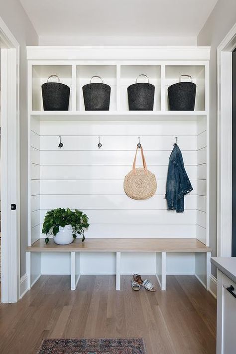 This welcoming mudroom features a light wooden bench fixed against a shiplap backsplash beneath coat hooks mounted under white cubbies holding black baskets. Shiplap Wall Storage, Entry Way Built In Bench With Storage, Built In Bench Open Underneath, Custom Entry Bench, Mudroom Seating Ideas, Kitchen Nook Mudroom, Open Bench Seating, White Oak Mudroom Bench, Entry Way Bench Built In