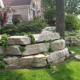 Natural Ledgerock Outcropping Stone was used to create a natural retaining wall to support the fireplace patio and preserve the mature oak tree. Perennial Plantings were installed within the retaining wall to add color and soften the appearance. Tree Wall Landscaping, Large Boulder Retaining Wall, Outcropping Retaining Wall, Large Stone Retaining Wall, Outcropping Landscaping, Front Yard Retaining Wall Curb Appeal, Limestone Retaining Wall Ideas, Retention Wall Ideas, Natural Retaining Wall