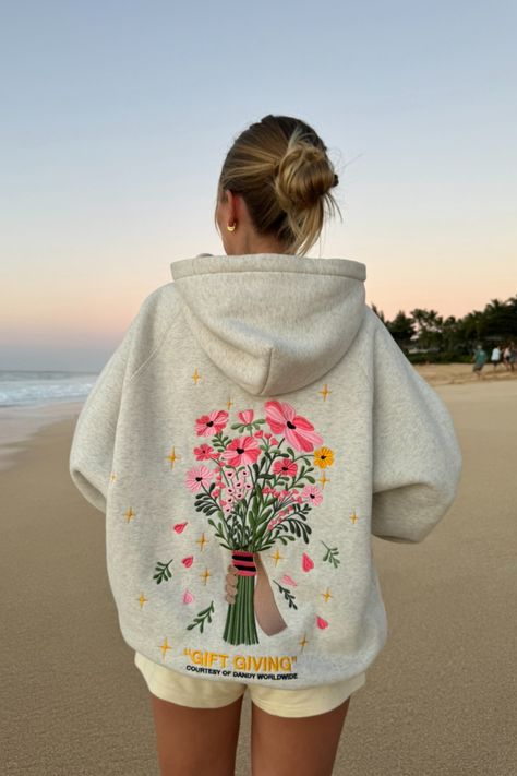 photo shows a blonde teenage girl standing on a sandy beach at sunset. she is facing away from the camera, looking at the ocean in the distance. she is wearing cream colored sweat shorts and a gray hoodie that has a big embroidered graphic of a bouquet of flowers on the back. "gift giving" and "courtesy of dandy worldwide" are embroidered on the back as well. the girl has her hair in a bun. the sky is orange and blue, like the sun is just about to set Dandy Flower Hoodie, Dandy Hoodie Gift Giving, Aesthetic Hoodies For Women, Gift Giving Hoodie, Danny Worldwide Hoodie, Cute Aesthetic Hoodies, Hoodies That Hoodie, Embroidery Designs On Hoodies, Dandy Worldwide Hoodie