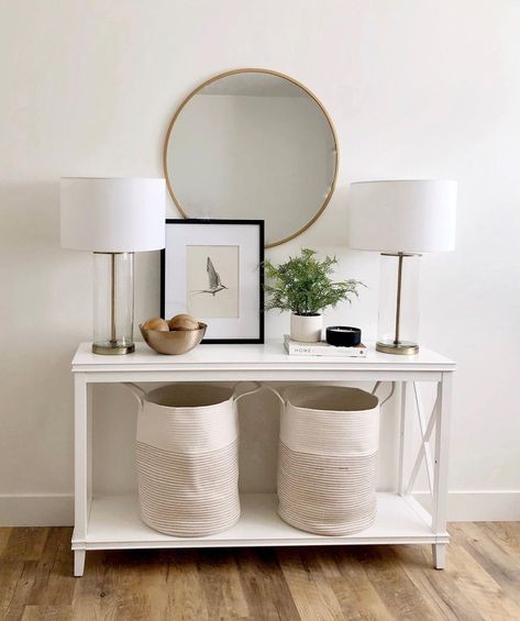 This bright hallway includes two tall class cylindrical lamps with white lampshades sitting on a white console table. A gold round lamp hangs on the white wall behind a simple picture framed in black. A potted plant next to a black bowl sits on a book. Two large cloth baskets rest underneath the console table. A warm wood floor runs the length of the hallway. Decorative Hamper, Blanket Basket For Living Room, White Entryway Table, Country Girl Home, Basket For Living Room, Basket Blanket, Blanket Holder, White Hallway, Warm Wood Flooring