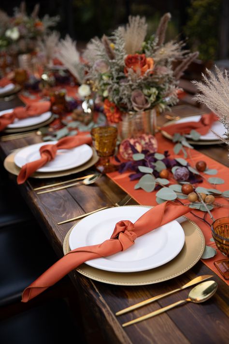 This beautiful tablescape is perfectly fall themed. Dried eucalyptus trails down the table runner which coordinates with the orange napkins on each plate. A gold charger and flatware ties in to the vases of the beautiful orange and dusty lilac flowers. Pampas grass adds a fall touch and vintage style brown glasses add some extra interest. Seasonal fruits add a natural element to the table. Rust Orange Table Setting, Round Table Wedding Decor Burnt Orange, Brown And Orange Table Setting, Orange Theme Aesthetic, Fall Wedding Plate Setting, Dried Orange Place Setting, Gold Charger Plates Table Setting, Fall Chargers Plates, Burnt Orange Table Setting