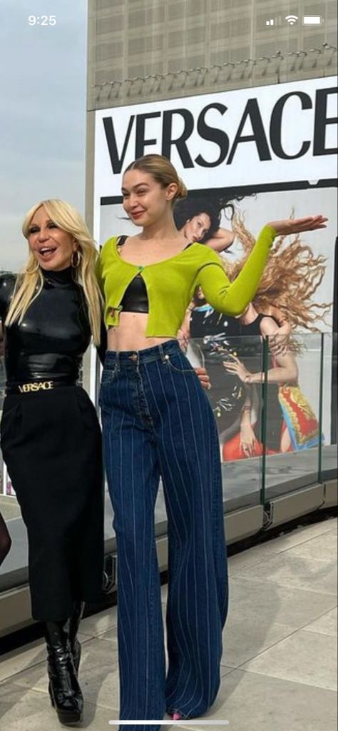 Gigi wearing a green sweater (with the length of a top) over a black top, she is also wearing dark blue jeans with white skinny vertical lines, her hair is in a middle bun and she is posing next to Donatella Versace who’s wearing a black leather o latex turtle neck-long-sleeved shirt, a black long tube skirt and a Versace black belt. They ate both in front of a Versace billboard for their most recent 2022 campaign. Gigi Hadid Outfits Casual, Gigi Hadid Off Duty, Gigi Hadid Casual, Gigi Hadid Photoshoot, Gigi Hadid Street Style, Gigi Hadid Outfits, Gigi Hadid Style, Fashion Model Poses, Hadid Style