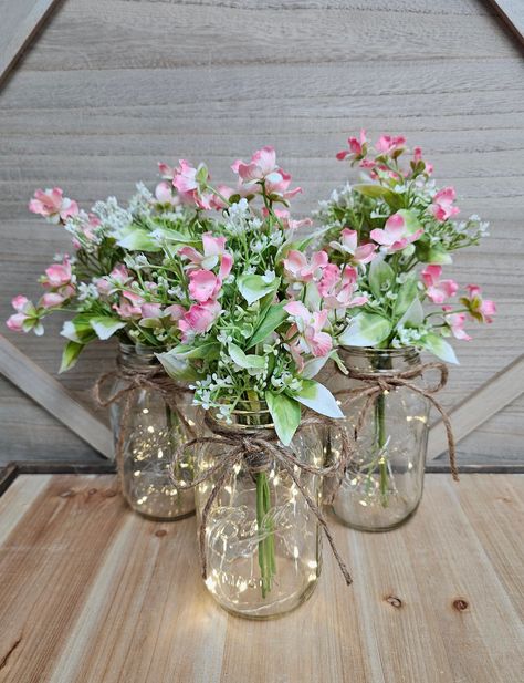 These beautiful faux wildflower and eucalyptus quart size mason jar centerpieces are a Glassy Gal original design. Each centerpiece includes one set of warm white LED fairy lights, a jute bow, and a deluxe faux wildflower and eucalyptus arrangement.  These centerpieces are very popular for weddings, baby showers, parties, corporate events, holiday events, and any other event that requires table centerpieces. They also look great in any room of your house or office. To see other lighted jar options and all of my other listings, please view my shop: https://www.etsy.com/shop/GlassyGalDecor  Each jar set includes: (1) clear quart size mason jar (1) set of LED fairy lights (1) faux wildflower and eucalyptus arrangement  (1) jute bow Dimensions:  Quart jars are approximately 6.25" tall (approxi Fairy Birthday Party Centerpieces, Pink And Green Centerpieces, Eucalyptus Arrangement, Jar Centerpiece Wedding, Water Centerpieces, Diy Floral Centerpieces, Romantic Table Decor, Sweet 16 Centerpieces, Fairy Baby Showers