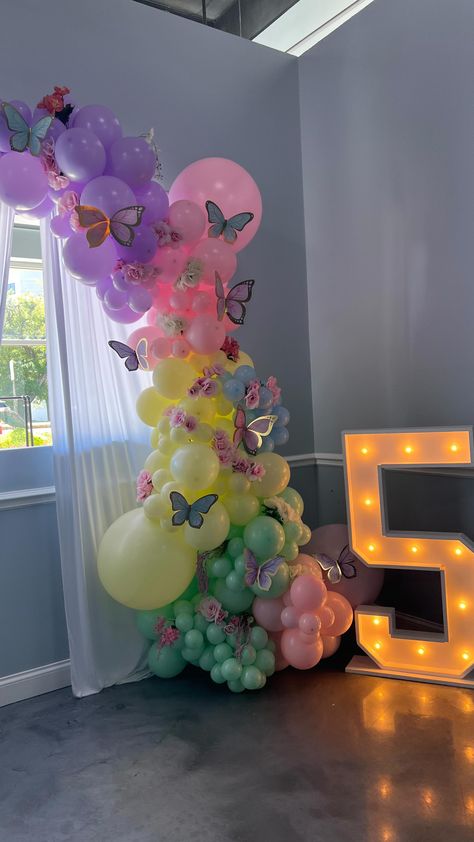 10 foot color blocked balloon garland with silk flowers and paper butterflies next to a marquee number 5 Butterfly And Flower Balloon Arch, Butterfly And Rainbow Birthday Party, Balloon Garland With Flowers And Butterflies, Flowers And Butterflies Decorations, Fairy Theme Balloon Decoration, Pastel Butterfly Balloon Garland, Unicorns And Butterflies Party, Butterfly Balloon Decorations, Birthday Theme Butterfly