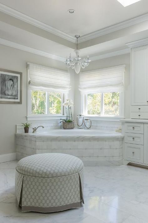 Beautiful master bathroom boasts a corner marble tiled tub placed under windows dressed in white roman shade illuminated by a white chandelier alongside a round skirted ottoman with grey ribbon trim. Jacuzzi Design, Tiled Tub, Bathroom Tub Remodel, Arabesque Tiles, Romantic Bathrooms, Beautiful Master Bathrooms, Tub Remodel, Bathroom Window Treatments, Bathtub Remodel