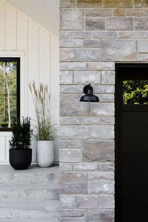 Black garage door. Farmhouse gooseneck sconce wall light. Stone and white board and batten exterior. Board And Batten Farmhouse, White And Black House Exterior, White Siding Exterior, Brick And Siding Exterior, White Board And Batten, White Farmhouse Exterior, Board And Batten Exterior, Stone Exterior Houses, House Colors Exterior