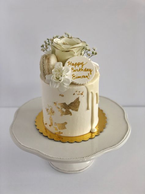 White cake with gold leaf details in front. White drip. White florals and macaron on top. There is a plaque that reads “Happy Birthday Simran!”. White Cake With Gold Flakes, Gold And White Cake Birthday, White And Gold Cake Ideas, White And Golden Cake, Engagement Desserts, White And Gold Cake, Gold And White Cake, Golden Birthday Cakes, Salon Party