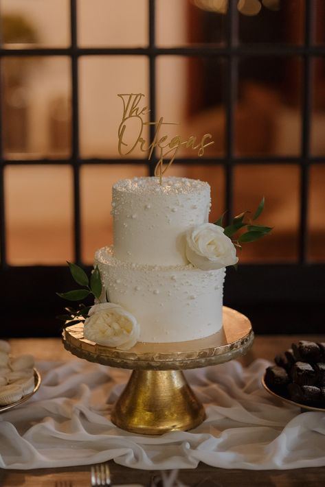 White wedding cake sits atop a gold wedding cake stand. The couple's last name is a decoration at the top of this timeless wedding cake Ivory Wedding Cake Elegant, Italian Style Wedding Cake, Elegant Simple Wedding Cake, Simple Classic Wedding Cake, Modern Romantic Wedding Cake, Wedding Cake Elegant Classy, Minimalistic Wedding Cake, Timeless Wedding Cake, Small Elegant Wedding