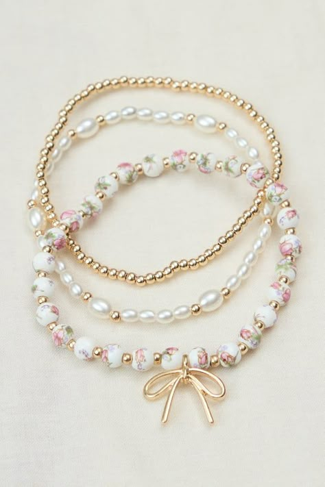 This elegant 3-pack of stretch bracelets adds a touch of sophistication to any outfit. Featuring gold beads, white pearl-inspired beads, and painted beads with a charming gold bow charm, this set offers versatile styling options for a polished look. Painted Beads, Preppy Jewelry, Pretty Jewelry Necklaces, Bow Bracelet, Diy Charm Bracelet, Beads Bracelet Design, Jewelry Accessories Ideas, Gold Bead Bracelets, Christmas Bracelet