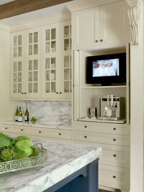 Glass front ivory kitchen cabinets are mounted above ivory shaker cabinets topped with a gray and white marble countertop placed against a matching backsplash and beside pull out shelves holding a TV and a coffee maker positioned beneath ivory cabinets. Small Kitchen Tv, Tv In The Kitchen, Ivory Kitchen Cabinets, Tv Kitchen, Ivory Kitchen, Glass Kitchen Cabinet Doors, Glass Kitchen Cabinets, Tv In Kitchen, Kitchen Tv