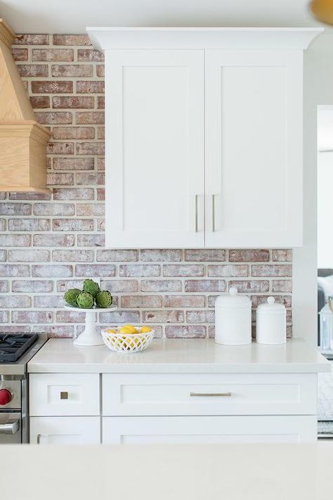 White shaker kitchen cabinets feature nickel knobs, white quartz countertops, and red brick backsplash tiles. White Kitchen With Brick, Red Brick Kitchen, Kitchen With Brick, Brick Kitchen Backsplash, Backsplash Kitchen White Cabinets, Brick Backsplash Kitchen, Rustic Backsplash, White Shaker Kitchen Cabinets, White Shaker Kitchen