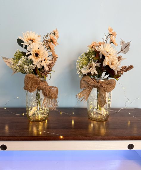 Are you looking for a unique and charming way to brighten up your space? Whether you are planning a wedding, an engagement, or any other special occasion, or you just want to add some cozy and rustic vibes to your home, these neutral earth tone wildflower centerpieces are perfect for you! Each centerpiece features a beautiful bouquet of hand-picked artificial wildflowers in a Mason jar with lights and a burlap bow. The flowers are carefully arranged to create a natural and harmonious look, and the lights add a touch of warmth and magic. The burlap bow adds a finishing touch of elegance and simplicity. These centerpieces are available in either pint size or quart size, and you can choose from sets of 2, 4, 6, or 8. They are easy to display and move around, and they will make any room feel m Mason Jar With Burlap Ribbon, Mason Jar Western Centerpieces, Burgundy Mason Jar Centerpieces, Mason Jar Fairy Lights Centerpieces, Mason Jar Fall Flower Arrangements, Rustic Centerpieces For Party, Rustic Baby Shower Centerpieces, Growler Centerpiece Wedding, Rustic Wedding Centerpieces Diy