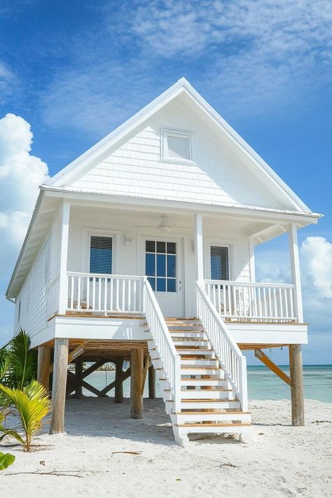 Tiny cottage style beachfront house on stilts with stairs leading to the entry clapboard siding. Discover tiny beach houses that are big on charm and functionality. You’ll want to own one. Raised Beach House Plans, Small Beach Bungalow, Tiny House Beach, House On Piers, Narrow Beach House, Beach House Plans On Stilts, Cottage On Stilts, Beach Shack Exterior, Beach Side House