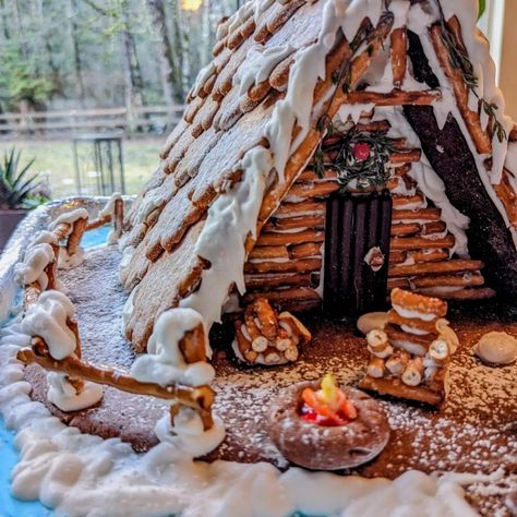 Gingerbread a-frame cabin on a lake with a candy campfire and mini chair and woodpile. To the side, a mini snowman and rosemary tree. The entrance to the pretzel log cabin is adorned with a thyme and candy wreath, and a jelly bean path. Pretzel Cabin Christmas, Mountain Gingerbread House, Pretzel Gingerbread House Log Cabins, Non Gingerbread House Ideas, Ginger Bread Ideas Creative, Triangle Gingerbread House Ideas, Log Cabin Pretzel House, Lake House Gingerbread House, Pretzel House Log Cabins