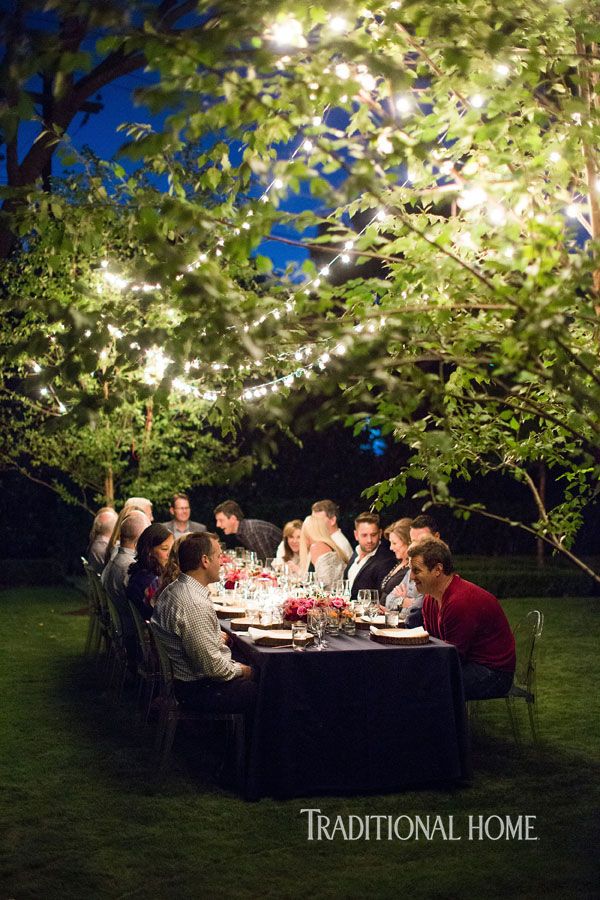 a group of people sitting around a dinner table under trees with lights strung from them