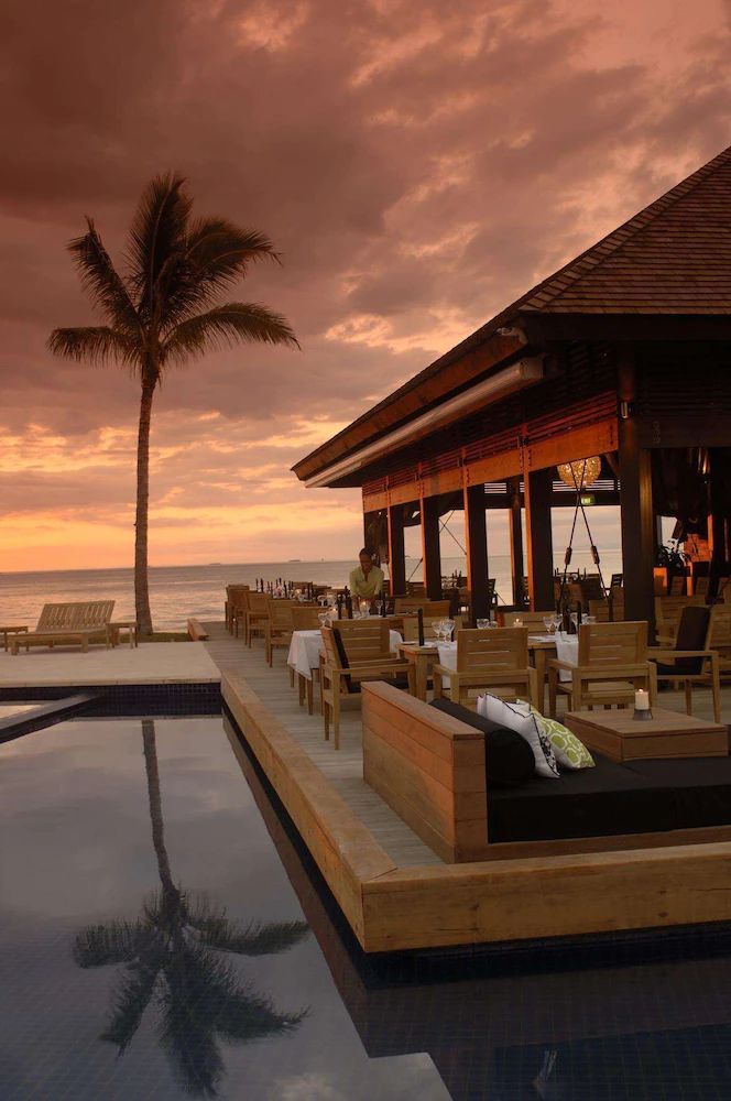 an outdoor dining area next to the ocean with palm trees in the background at sunset