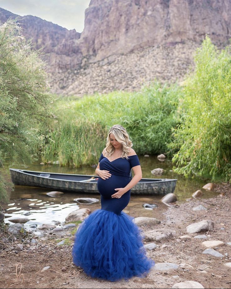 a pregnant woman standing in front of a river with a canoe on the other side