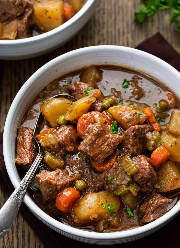 two white bowls filled with beef stew on top of a wooden table and garnished with parsley