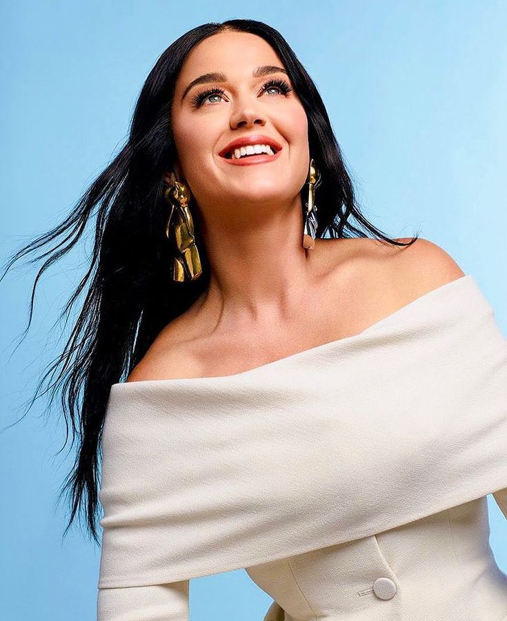 a woman with long black hair wearing a white top and gold earrings, posing for the camera