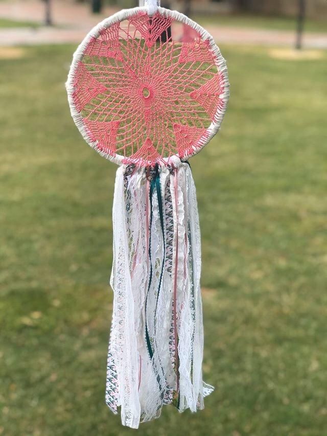 a pink and white dream catcher hanging from a string in the grass with trees in the background