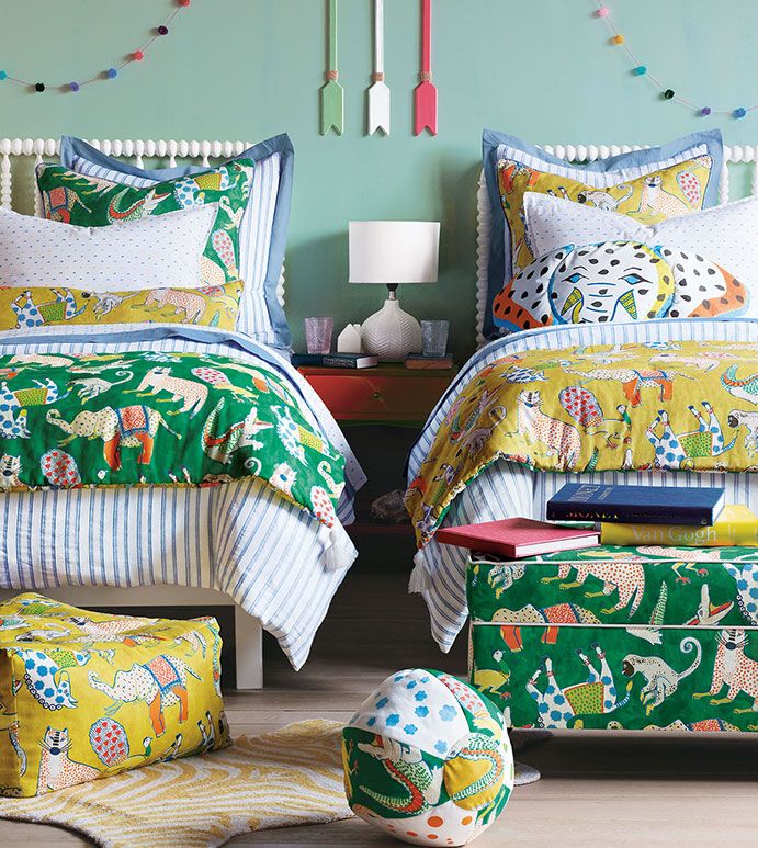 two beds with colorful comforters and pillows in front of a blue wall, one has a yellow footstool