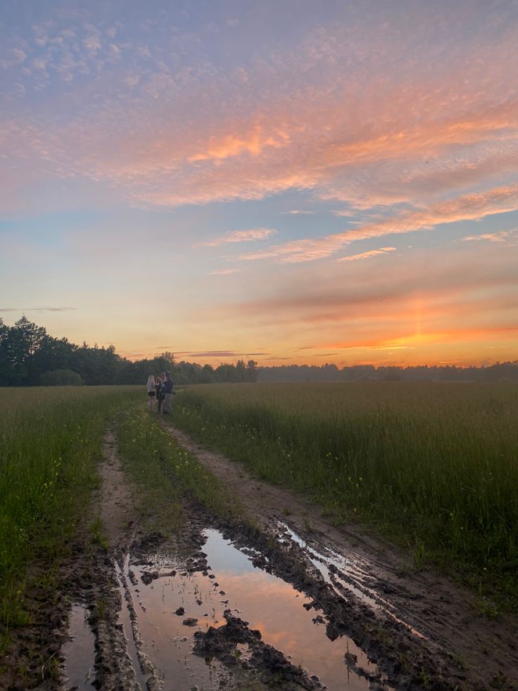 two people walking down a dirt road at sunset with puddles in the mud on either side