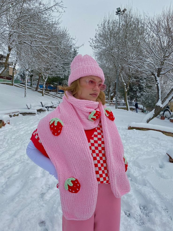 a woman standing in the snow wearing a pink knitted scarf and hat with strawberries on it