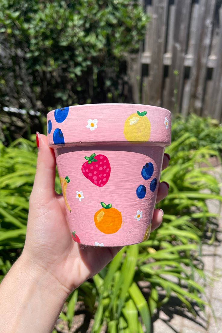 a hand holding up a pink cup with fruit on it and flowers in the background