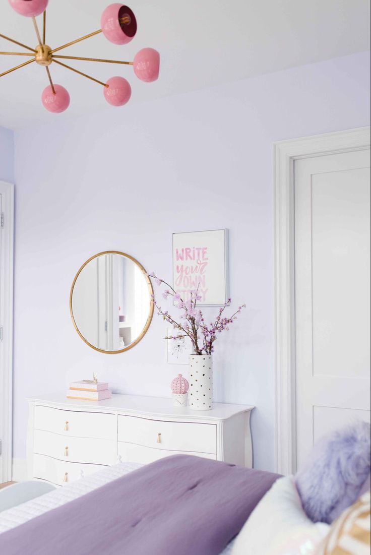 a bedroom decorated in pastel colors with pink and purple accents on the walls, white dressers, and a round mirror