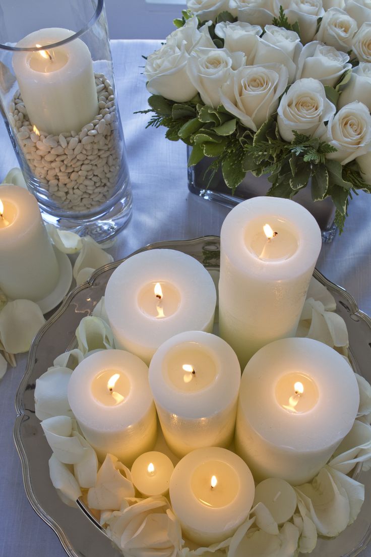 white candles are arranged in a bowl on a table with flowers and glass vases