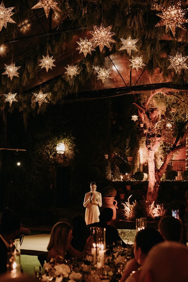 a woman standing in front of a crowd at a dinner table with lights on the ceiling