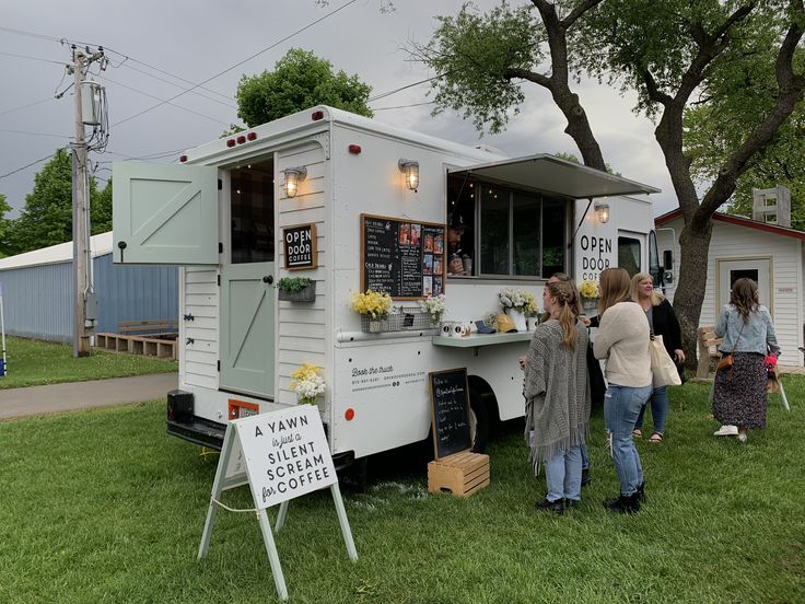 people are standing in front of a food truck on the grass near some trees and buildings