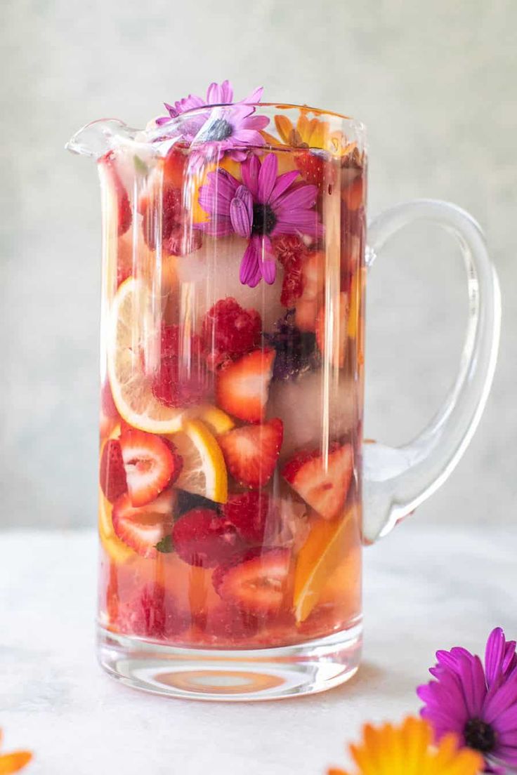 a pitcher filled with fruit and flowers sitting on top of a white table next to purple daisies