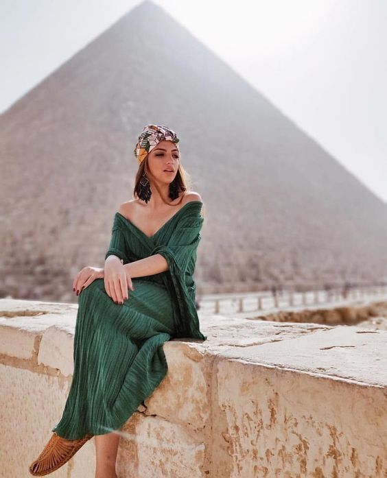 a woman in a green dress is sitting on a ledge near the pyramid of giza