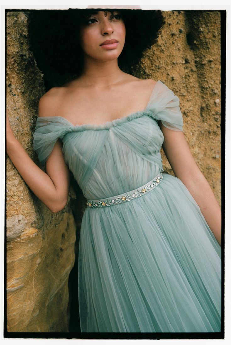 a woman in a blue dress leaning against a stone wall with her hand on her hip