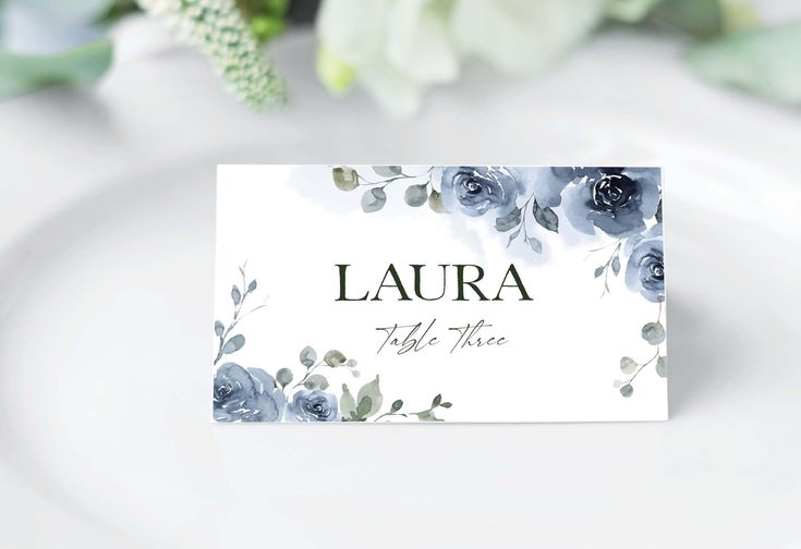 a place card with blue flowers on it is sitting on a white plate next to some greenery