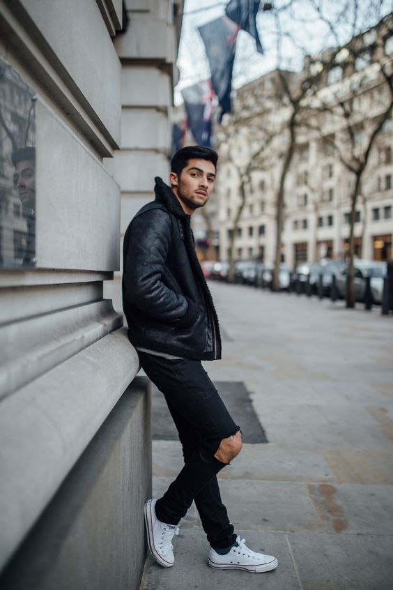 a man leaning against the side of a building wearing a black jacket and white sneakers