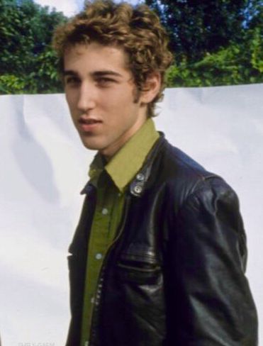 a young man wearing a leather jacket and green shirt standing in front of a white wall