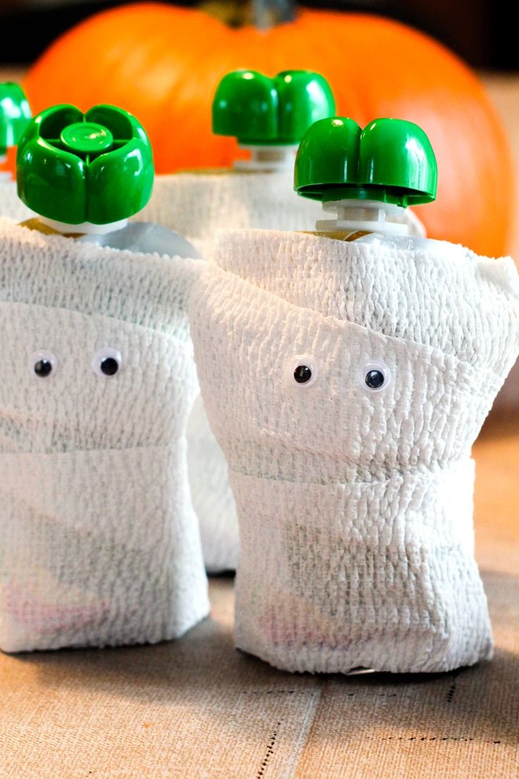 two baby bottles wrapped in white towels with green caps and eyes on them, sitting next to pumpkins