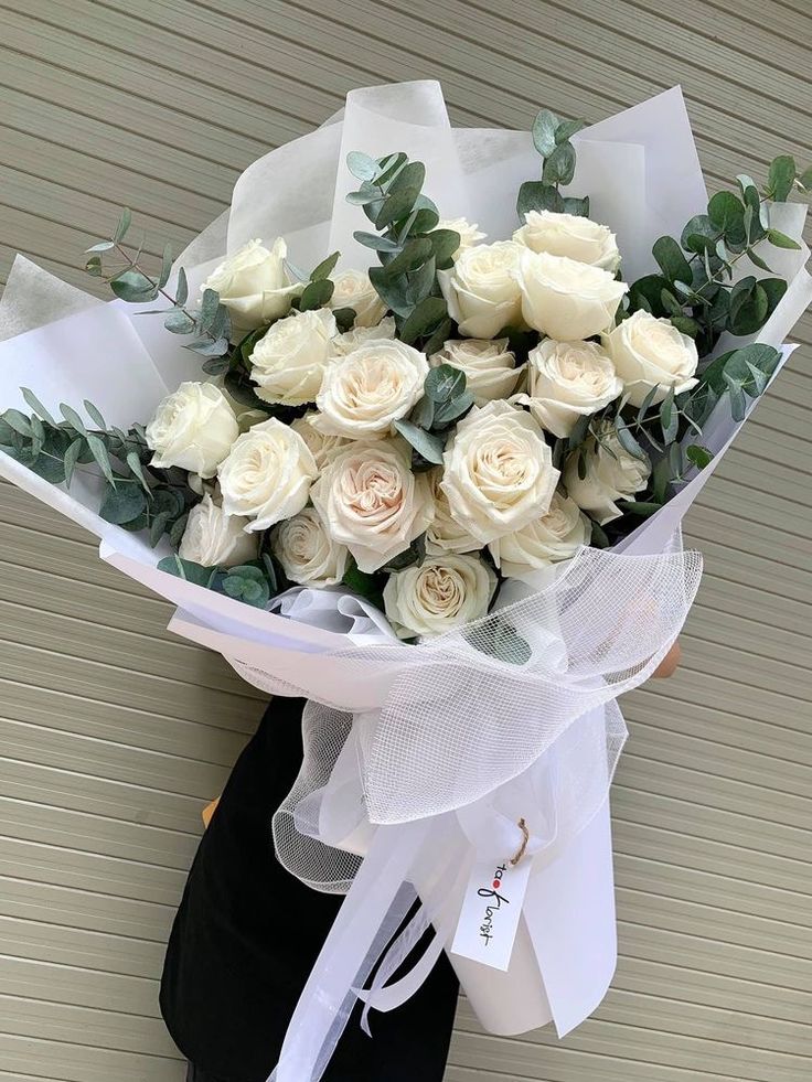 a bouquet of white roses with greenery wrapped around it, in front of a wall