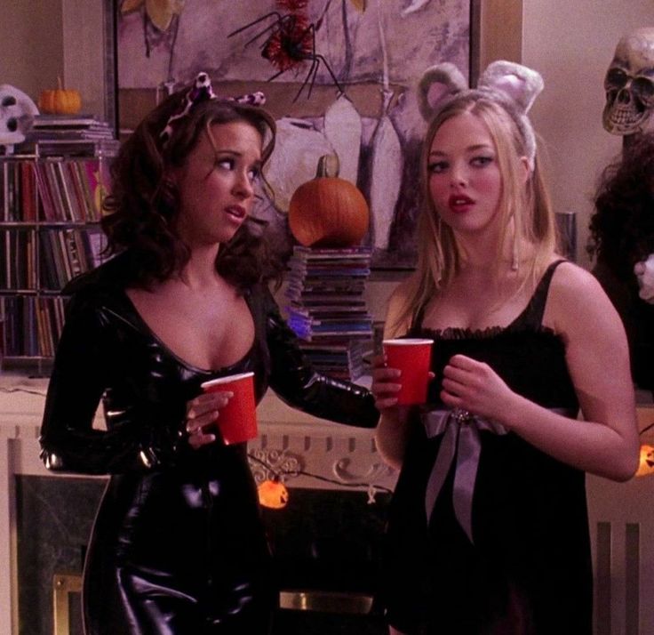 two women dressed up in costumes holding cups and looking at each other with halloween decorations on the wall behind them