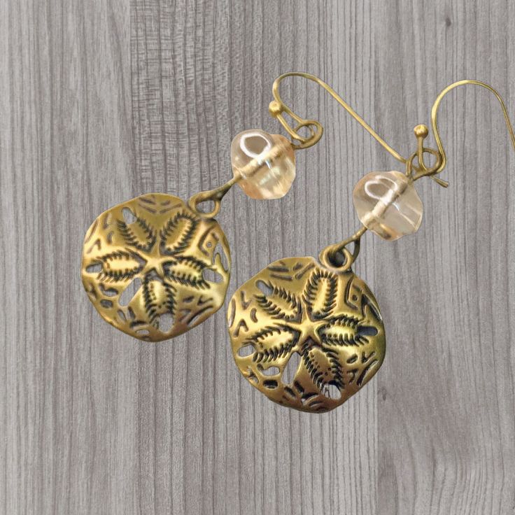 Brass sand dollar charms dangle playfully from champagne colored snail-shaped Czech glass beads. These earrings will add a touch of ocean vibe to any outfit and remind you of the beach. Sand dollars,  shell shaped beads, the color of sand, offer an elegant ocean vibe. The neutral colors beautifully comliment your natural clothing and can be worn with any color outfit. Artisan glass beads produced in small quantities in Czech Republic  Hypoallergenic ear wires (nickel and lead free). Select ear w Metal Beaded Earrings For Beach, Beach Beaded Metal Earrings, Beach Brass Dangle Earrings, Metal Beaded Drop Earrings For Beach, Beaded Metal Drop Earrings For Beach, Gold Beaded Dangling Earrings For Beach, Gold Dangling Beads Earrings For Beach, Round Metal Earrings For The Beach, Round Metal Earrings For Beach