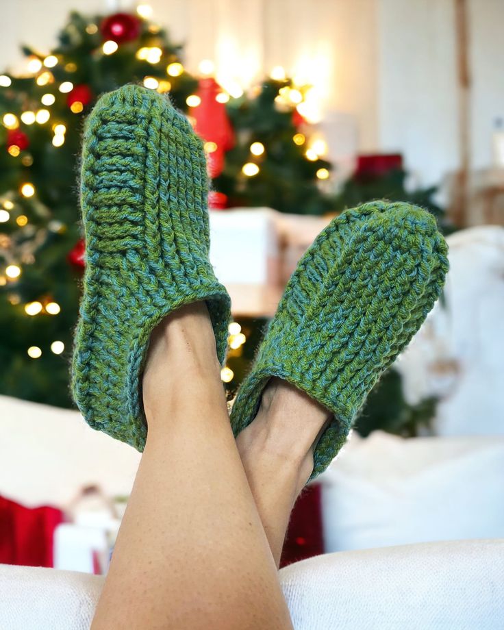 a woman's feet wearing green knitted slippers in front of a christmas tree