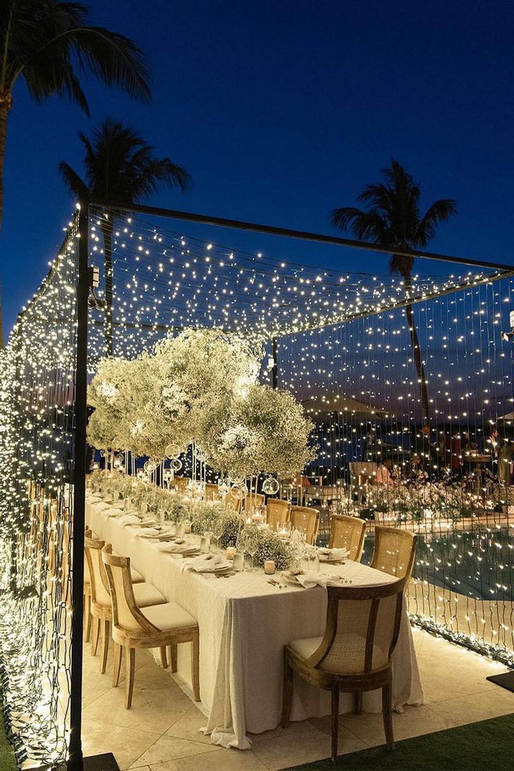 an outdoor dining table set up with lights and greenery on the side at night