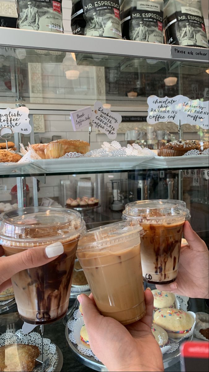 two people holding up cups of coffee in front of a display case with pastries