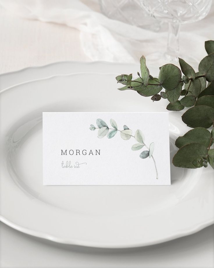 a white plate with a place card on it next to some green leaves and a glass vase